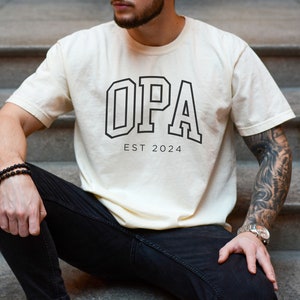 May include: A cream colored t-shirt with a black graphic that says "OPA EST 2024".