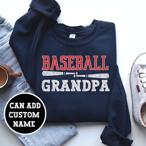 Peut inclure: Sweat-shirt bleu marine avec l'inscription "BASEBALL GRANDPA" en rouge et blanc. Le motif comprend un dessin de batte de baseball. Un cercle en bas à gauche indique "CAN ADD CUSTOM NAME".