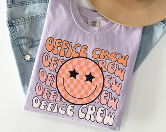 May include: A lilac-colored t-shirt with the words "OFFICE CREW" in orange and pink, along with a smiley face graphic. The shirt is folded on top of blue jeans, a gold necklace, and a tan hat.
