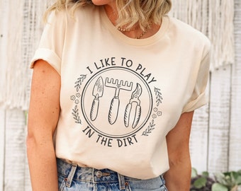 May include: A beige t-shirt with a black graphic design that says "I like to play in the dirt" with a circle around the text and three gardening tools inside the circle.