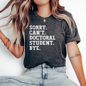 May include: A dark gray t-shirt with the text "SORRY. CAN'T. DOCTORAL STUDENT. BYE." printed in white.
