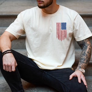 May include: A person wearing a cream colored t-shirt with a distressed American flag design on the left side of the chest. The person is wearing black jeans and a black beaded bracelet.