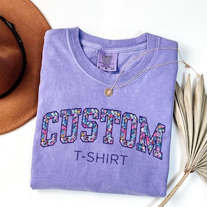 May include: A lavender Comfort Colors t-shirt with the word "CUSTOM" in a floral design. The text "T-SHIRT" is below the word "CUSTOM". A gold necklace with a pendant is on the shirt.