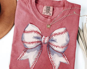 May include: A red t-shirt with a white baseball-stitched bow on the front. The shirt has the text "COMFORT COLORS" and "S - P - CH" on the tag.