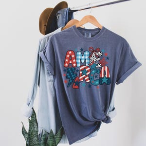May include: A blue t-shirt with the word "AMERICA" in a colourful, patriotic design. The letters feature patterns like stars, stripes, and plaid in red, white, and blue. The shirt is hanging on a wooden hanger.