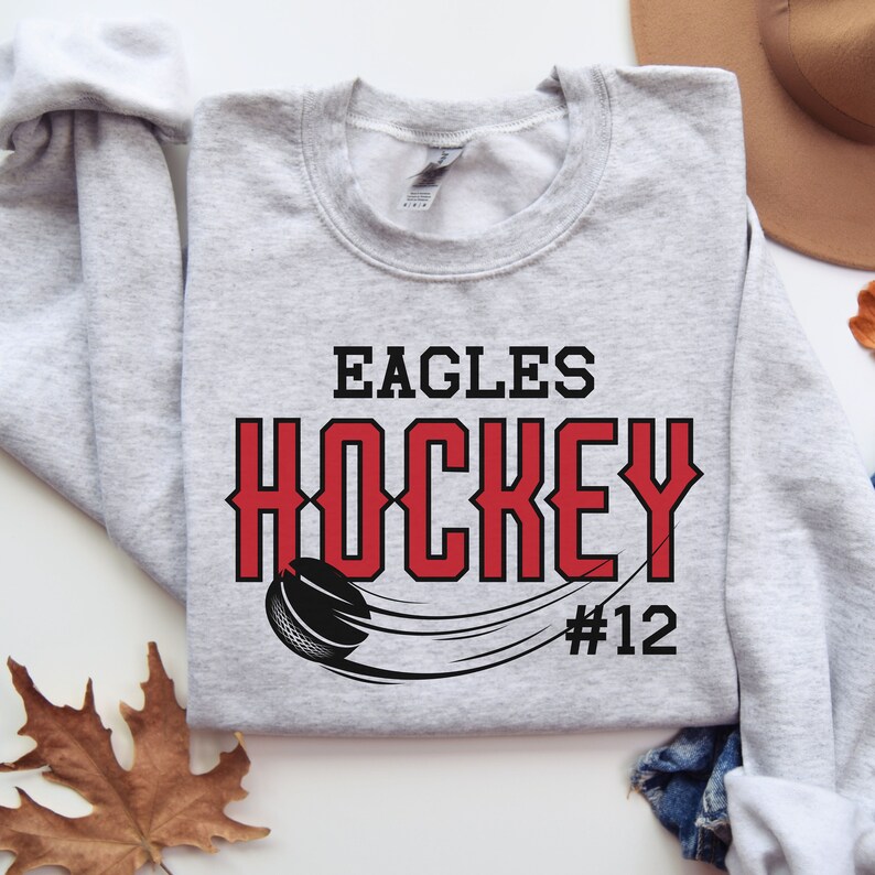 K&ouml;nnte beinhalten: Grauer Sweatshirt mit dem Text "EAGLES HOCKEY #12" in schwarzer und roter Schrift. Unter dem Text befindet sich eine Schwarzwei&szlig;illustration eines Hockeyschl&auml;gers und Pucks.