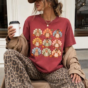 May include: A brick red short-sleeved crew neck t-shirt featuring a grid of nine stylized turkey illustrations in various patterns and colors, including blue, orange, and green, accented with small autumn leaves on a solid red background.
