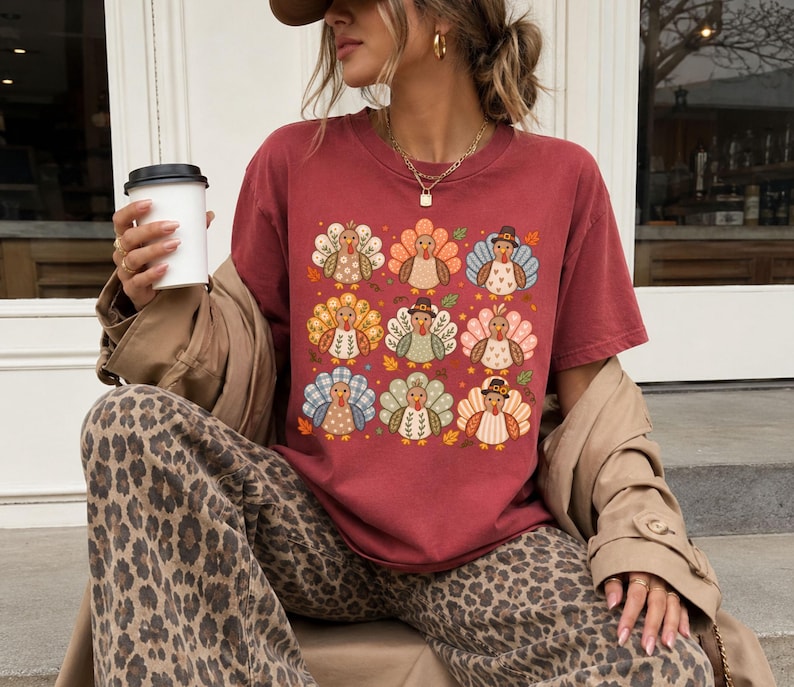 May include: A brick red short-sleeved crew neck t-shirt featuring a grid of nine stylized turkey illustrations in various patterns and colors, including blue, orange, and green, accented with small autumn leaves on a solid red background.