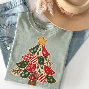 May include: A sage green t-shirt featuring a large, layered Christmas tree graphic made of various red, green, and cream fabric-style patches with patterns like plaid, polka dots, and snowflakes, topped with a gold star.
