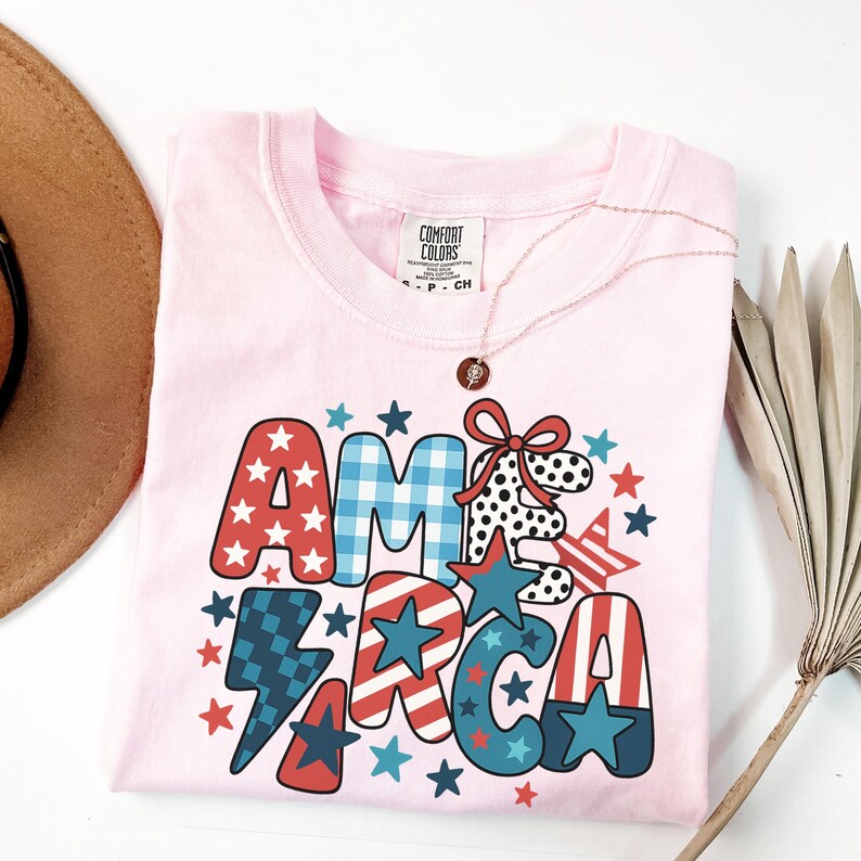 May include: A pale pink Comfort Colors t-shirt featuring the word "AMERICA" in a patriotic design. The letters are decorated with stars, stripes, and gingham patterns in red, white, and blue. A gold necklace with a pendant is draped on the shirt.