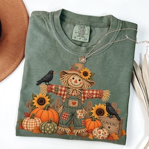 May include: A sage green short-sleeve t-shirt featuring a graphic of a scarecrow wearing a hat with a sunflower, standing near pumpkins, sunflowers, and two black crows. A wooden crate labelled PUMPKINS is visible. The shirt has a Comfort Colors label.