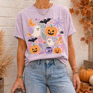 Może przedstawiać: Jasnofioletowy t-shirt z kr&oacute;tkim rękawem z kolorowym nadrukiem graficznym o tematyce Halloween. Wz&oacute;r zawiera pomarańczowe dynie, białe duszki, czarne nietoperze, fioletową pajęczynę z pająkiem, fioletowy kociołek oraz cukierki.