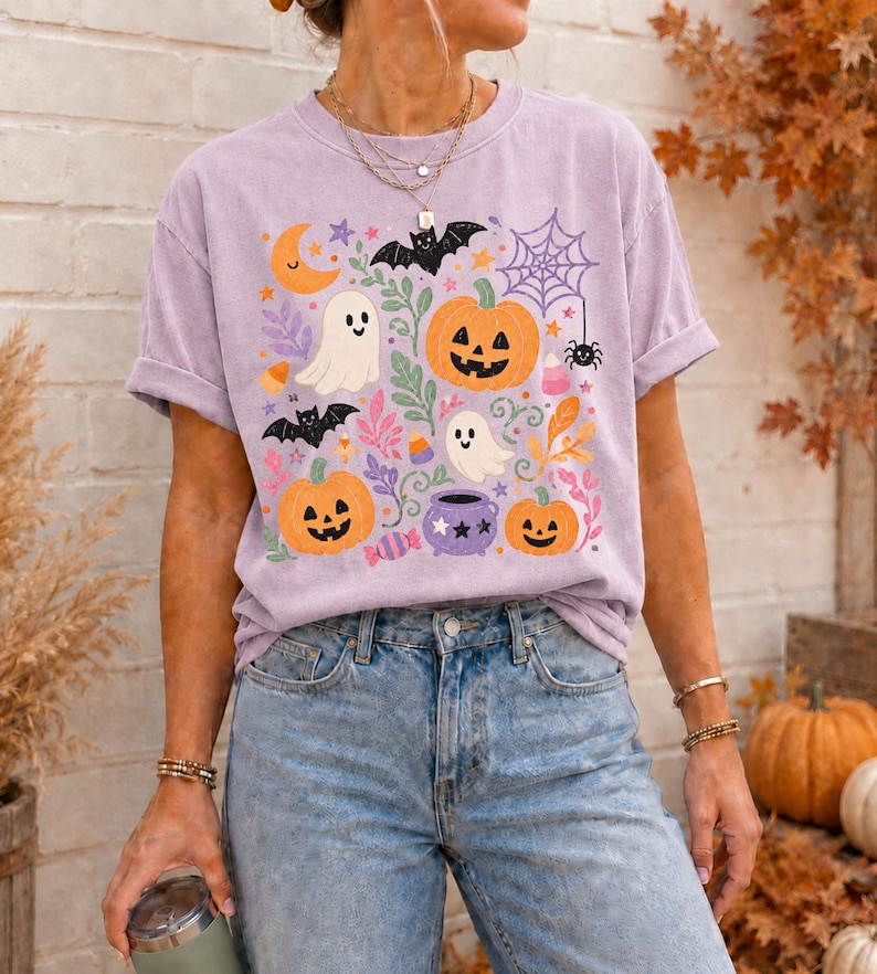 Może przedstawiać: Jasnofioletowy t-shirt z kr&oacute;tkim rękawem z kolorowym nadrukiem graficznym o tematyce Halloween. Wz&oacute;r zawiera pomarańczowe dynie, białe duszki, czarne nietoperze, fioletową pajęczynę z pająkiem, fioletowy kociołek oraz cukierki.