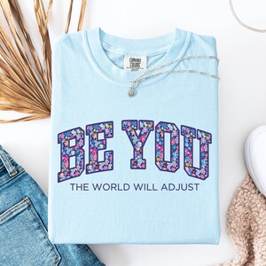Pode incluir: Uma t-shirt azul claro com o texto BE YOU numa fonte universit&aacute;ria preenchida com um padr&atilde;o floral colorido. Abaixo do texto principal, a frase THE WORLD WILL ADJUST est&aacute; impressa numa fonte azul escura simples. A t-shirt tem gola redonda.