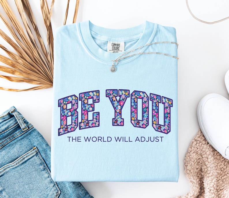 Pode incluir: Uma t-shirt azul claro com o texto BE YOU numa fonte universit&aacute;ria preenchida com um padr&atilde;o floral colorido. Abaixo do texto principal, a frase THE WORLD WILL ADJUST est&aacute; impressa numa fonte azul escura simples. A t-shirt tem gola redonda.