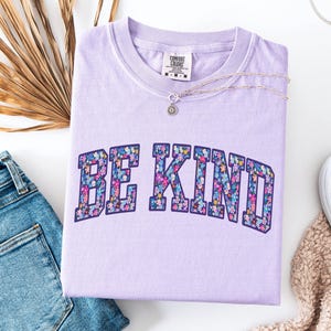May include: A light purple Comfort Colors t-shirt with the words "BE KIND" in a floral design. A silver necklace with a small pendant rests on the shirt. The shirt is paired with blue jeans and a beige scarf.