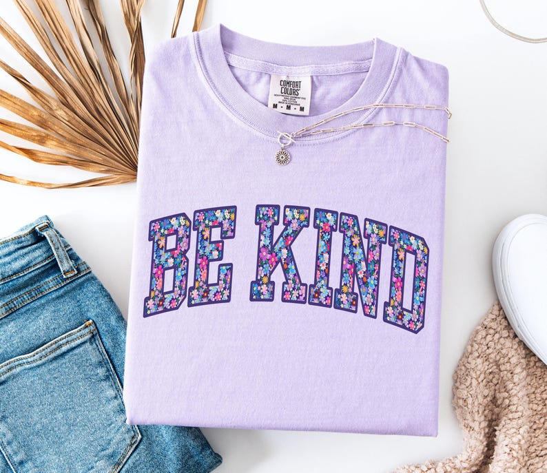 May include: A light purple Comfort Colors t-shirt with the words "BE KIND" in a floral design. A silver necklace with a small pendant rests on the shirt. The shirt is paired with blue jeans and a beige scarf.