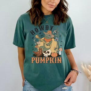 Pu&ograve; includere: Una t-shirt a maniche corte verde scuro con una stampa grafica di una rana che indossa un cappello da cowboy e una bandana, che suona il banjo seduta su un teschio. Il design include una zucca, funghi, fiori e la scritta HOWDY PUMPKIN.