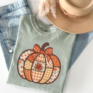May include: A pale green t-shirt with a pumpkin design. The pumpkin is made of patchwork patterns, including floral and gingham prints, and has a bow. A tan hat and blue jeans are also in the image.