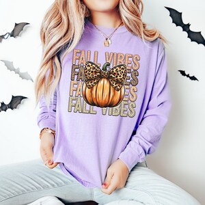 May include: A lilac long-sleeved top with a pumpkin design and the words "FALL VIBES". The pumpkin has a leopard print bow. The shirt is a casual style, suitable for autumn.