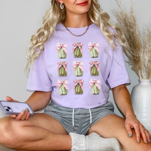 May include: A lavender t-shirt with a repeated pattern of pickle jars tied with pink bows. The jars are filled with green pickles.