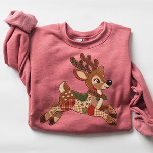 May include: A dusty rose long-sleeve crewneck sweatshirt featuring a large, central appliqué of a leaping reindeer. The reindeer is constructed from various fabric patches in shades of brown, green, and red, with a small holly sprig at its neck.