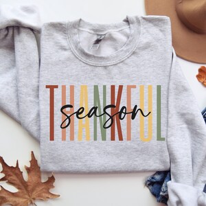 Puede incluir: Una sudadera gris con el texto "Thankful Season" en letras negras. El texto est&aacute; rodeado de rayas verticales en tonos naranja, amarillo, verde y marr&oacute;n.