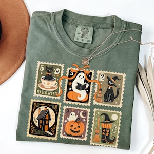 May include: Green Comfort Colors t-shirt with a Halloween-themed stamp design. The stamps feature a black cat, ghost, pumpkin, witch's hat, and haunted house. A silver necklace with a small pendant rests on the shirt.