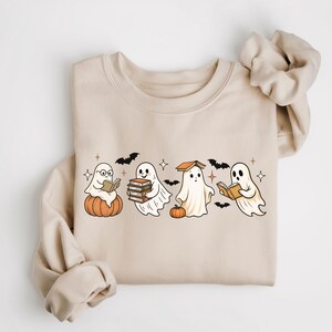 Puede incluir: Sudadera beige con un dise&ntilde;o de Halloween. El gr&aacute;fico muestra cuatro fantasmas de dibujos animados leyendo libros, con murci&eacute;lagos y estrellas. Un fantasma est&aacute; sentado en una calabaza. La sudadera tiene mangas largas y cuello redondo.