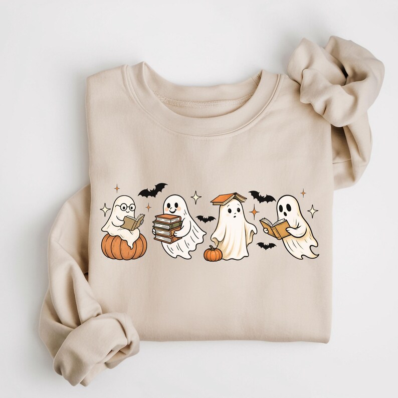 Puede incluir: Sudadera beige con un dise&ntilde;o de Halloween. El gr&aacute;fico muestra cuatro fantasmas de dibujos animados leyendo libros, con murci&eacute;lagos y estrellas. Un fantasma est&aacute; sentado en una calabaza. La sudadera tiene mangas largas y cuello redondo.