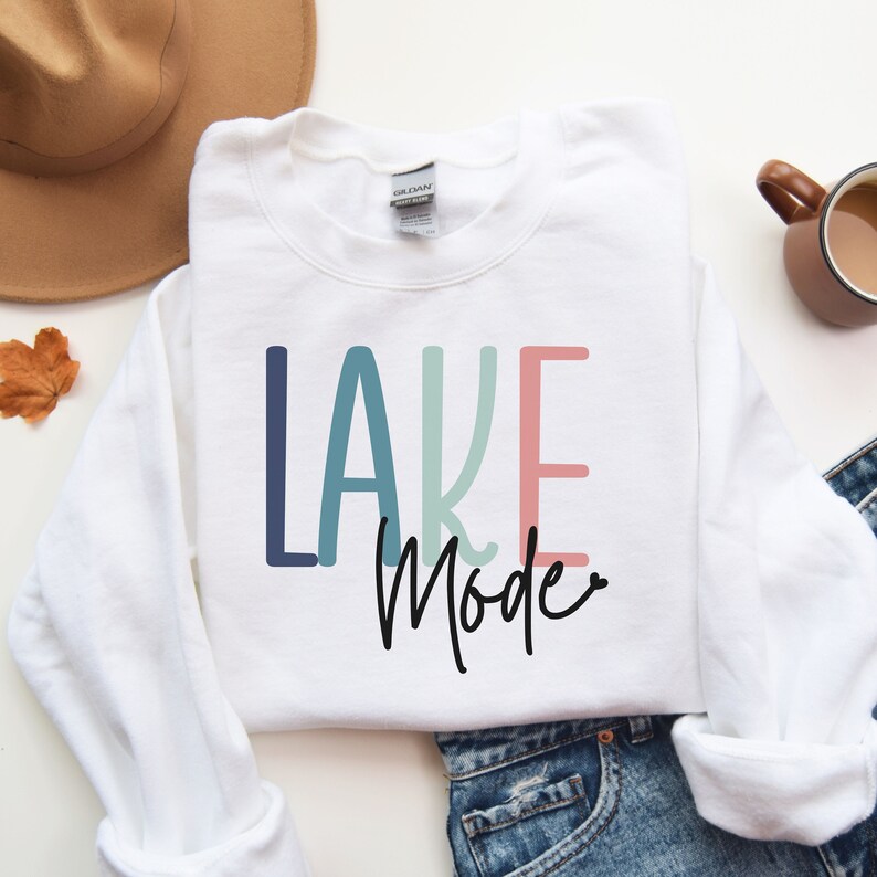 May include: White sweatshirt with the text "LAKE MODE" in colorful letters. The word "LAKE" is in blue, teal, and pink. The word "MODE" is in black cursive.