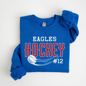 K&ouml;nnte beinhalten: Ein k&ouml;nigsblauer Pullover mit den Worten "Eagles Hockey #12" in wei&szlig;er und roter Schrift. Ein Hockeypuck und ein Schl&auml;ger sind in Wei&szlig; dargestellt.