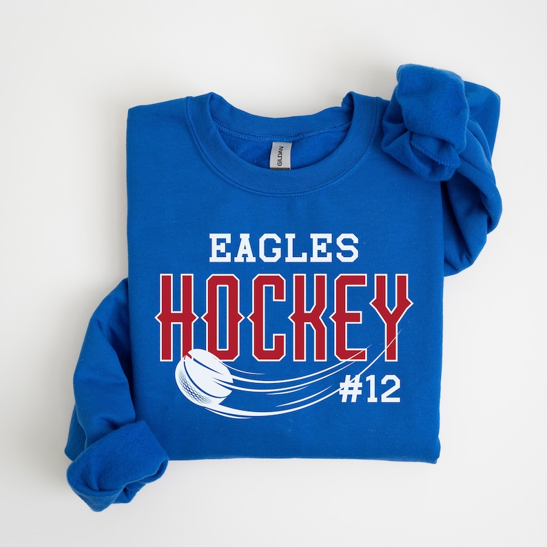 K&ouml;nnte beinhalten: Ein k&ouml;nigsblauer Pullover mit den Worten "Eagles Hockey #12" in wei&szlig;er und roter Schrift. Ein Hockeypuck und ein Schl&auml;ger sind in Wei&szlig; dargestellt.
