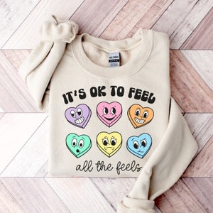 Pu&ograve; includere: Una felpa beige con la scritta "It's OK to Feel all the feels" stampata sul davanti. Il testo &egrave; in un font retr&ograve; ed &egrave; circondato da cuori colorati di cartoni animati con diverse espressioni facciali.