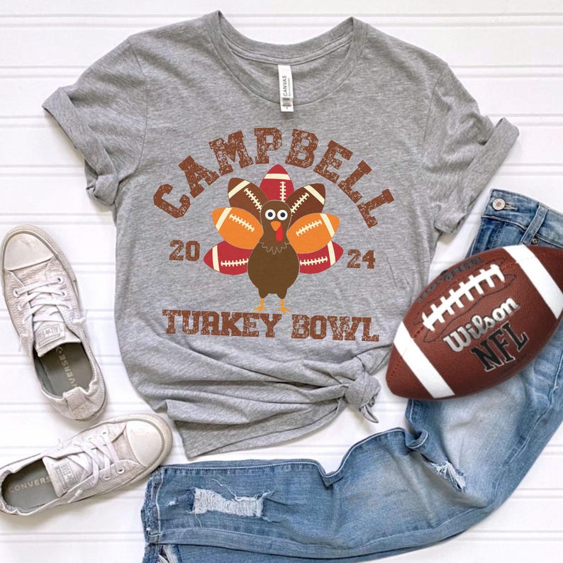 Op de afbeelding: Grijs T-shirt met een bruine kalkoen die een voetbalhelm draagt en voetballen vasthoudt. De tekst "Campbell 20 24 Turkey Bowl" staat op het shirt gedrukt.