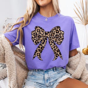 K&ouml;nnte beinhalten: Ein lila T-Shirt mit einer gro&szlig;en Schleife im Leopardenmuster auf der Vorderseite.