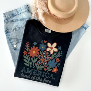 Peut inclure: T-shirt noir avec un motif floral compos&eacute; de fleurs rouges, blanches et bleues. Le texte "AMERICA. land of the free" est imprim&eacute; sous les fleurs.