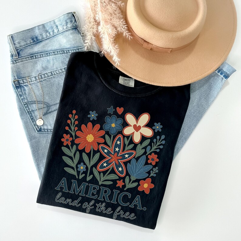 Peut inclure: T-shirt noir avec un motif floral compos&eacute; de fleurs rouges, blanches et bleues. Le texte "AMERICA. land of the free" est imprim&eacute; sous les fleurs.