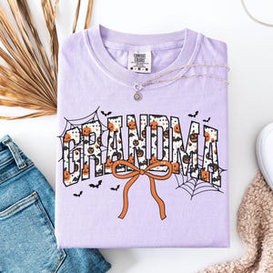 May include: A light purple t-shirt with the word "GRANDMA" in a Halloween-themed design. The letters are filled with pumpkins and bats, with an orange bow at the bottom. A silver necklace and a pair of jeans are next to the shirt.