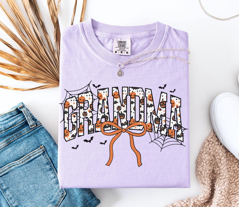 May include: A light purple t-shirt with the word "GRANDMA" in a Halloween-themed design. The letters are filled with pumpkins and bats, with an orange bow at the bottom. A silver necklace and a pair of jeans are next to the shirt.