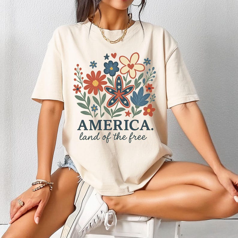 Peut inclure: Un t-shirt beige avec un motif de fleurs color&eacute;es et une &eacute;toile aux accents rouge, blanc et bleu. Le texte "AMERICA. land of the free" est imprim&eacute; sous le motif.