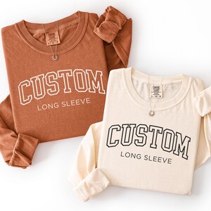 Op de afbeelding: Twee shirts met lange mouwen, &eacute;&eacute;n roestkleurig en &eacute;&eacute;n cr&egrave;mekleurig, worden getoond. Elk shirt heeft het woord "CUSTOM" in een collegiale lettertype, met "LONG SLEEVE" eronder. Beide shirts hebben een klein label en een ketting.