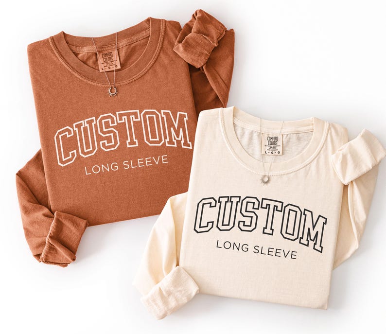 Op de afbeelding: Twee shirts met lange mouwen, &eacute;&eacute;n roestkleurig en &eacute;&eacute;n cr&egrave;mekleurig, worden getoond. Elk shirt heeft het woord "CUSTOM" in een collegiale lettertype, met "LONG SLEEVE" eronder. Beide shirts hebben een klein label en een ketting.