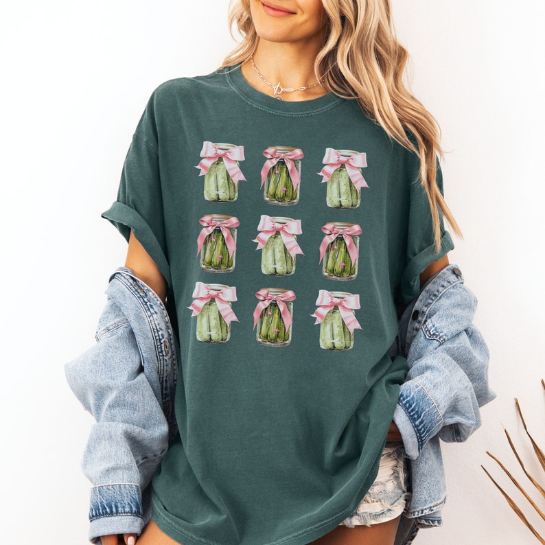 May include: A teal green t-shirt with a repeated pattern of nine jars of pickles tied with pink bows. Each jar contains green pickles.