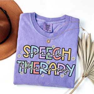 May include: A lavender-colored t-shirt with the words "SPEECH THERAPY" in a colorful, playful font. The design includes small daisy accents. A gold necklace with a pendant is draped across the shirt. The shirt is a Comfort Colors brand.