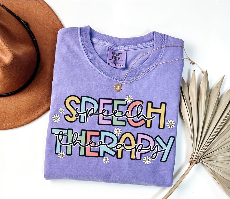 May include: A lavender-colored t-shirt with the words "SPEECH THERAPY" in a colorful, playful font. The design includes small daisy accents. A gold necklace with a pendant is draped across the shirt. The shirt is a Comfort Colors brand.