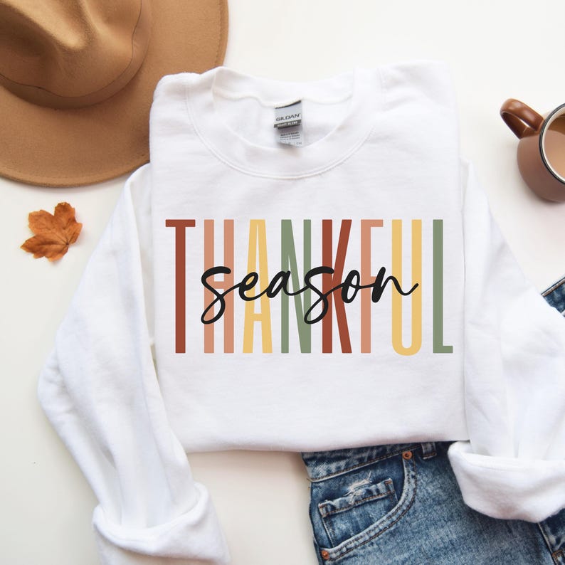Puede incluir: Sudadera blanca con el texto "Thankful Season" en rayas verticales de colores. El texto est&aacute; en un estilo de fuente moderno.