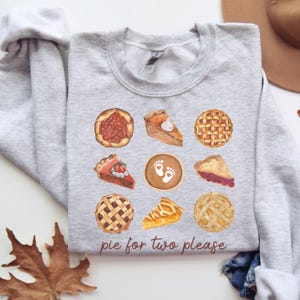 May include: A gray sweatshirt with a design of various pies, including cherry, pumpkin, apple, and pecan. The text "pie for two please" is printed in brown below the pies.