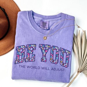 Pode incluir: Uma t-shirt de manga curta cor de lavanda com decote redondo. A parte da frente apresenta o texto BE YOU em letras grandes de bloco preenchidas com um padr&atilde;o floral colorido, seguido pelo texto THE WORLD WILL ADJUST em letras roxas.