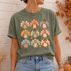 May include: An olive green t-shirt featuring a grid of nine stylized turkey illustrations. Each turkey has a unique patterned tail, including floral, plaid, and polka dot designs, with some wearing small hats, surrounded by scattered autumn leaves.